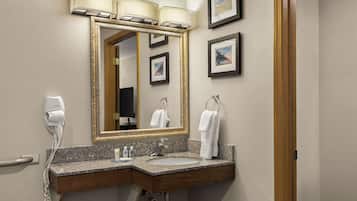 A hotel bathroom with a granite countertop sink, a large mirror with a gold frame, a hair dryer, and framed pictures on the wall.