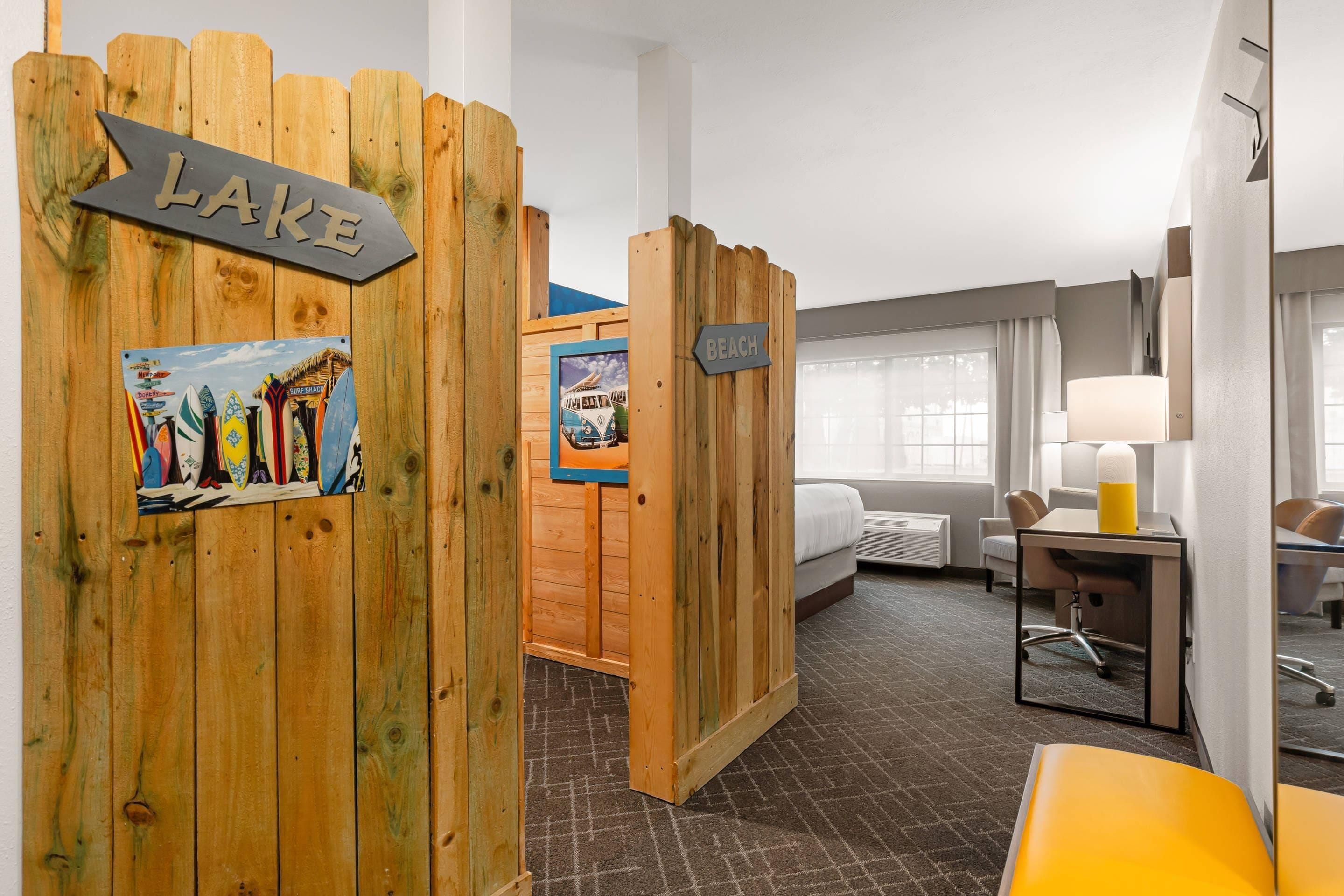 Une chambre d’hôtel avec une cloison en bois portant l’inscription « LAC », un lit, un bureau avec une chaise, une fenêtre avec des rideaux et une salle de bain avec une douche.