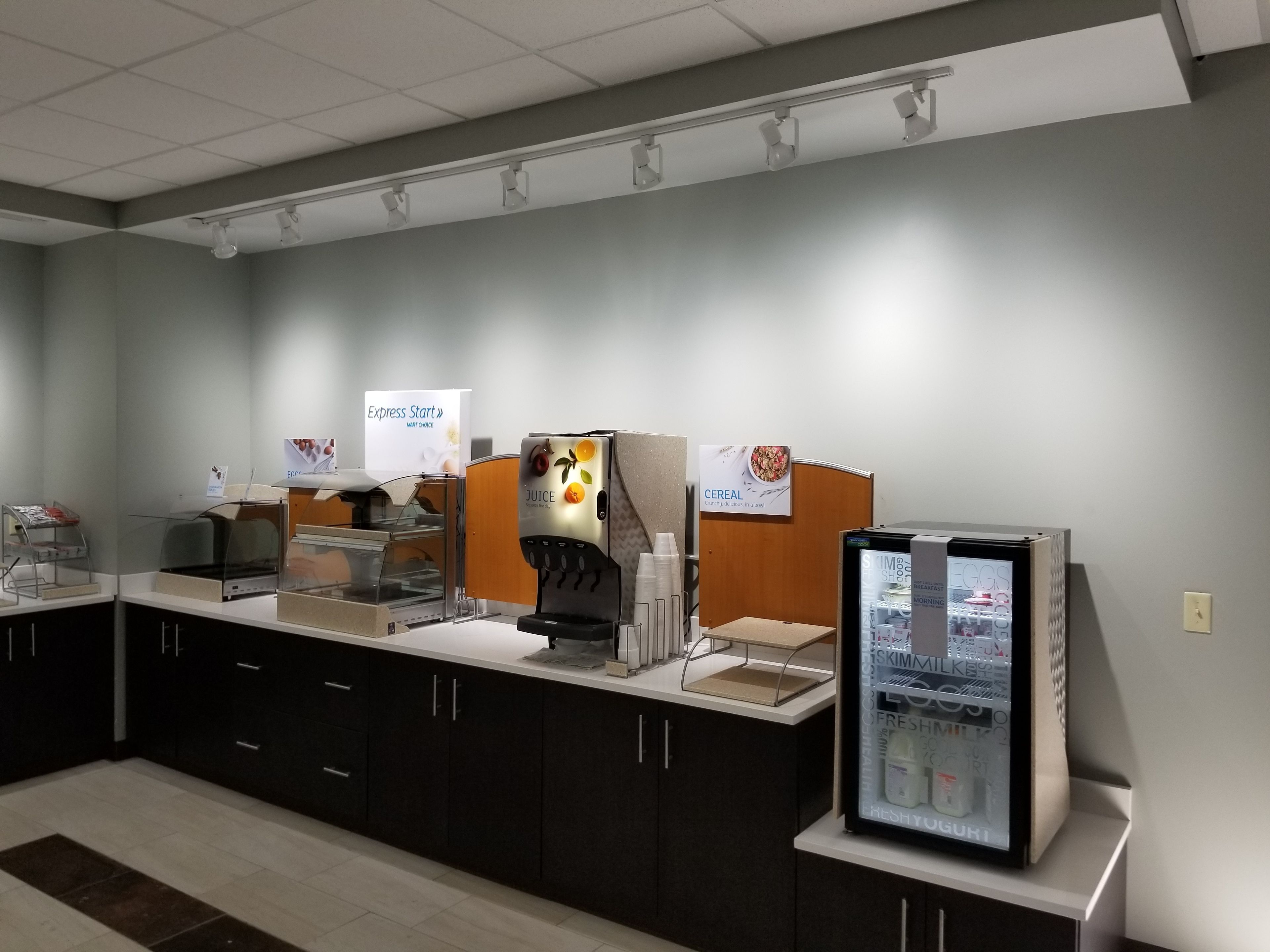 A self-service breakfast area with a juice dispenser, cereal station, and a refrigerated display case.