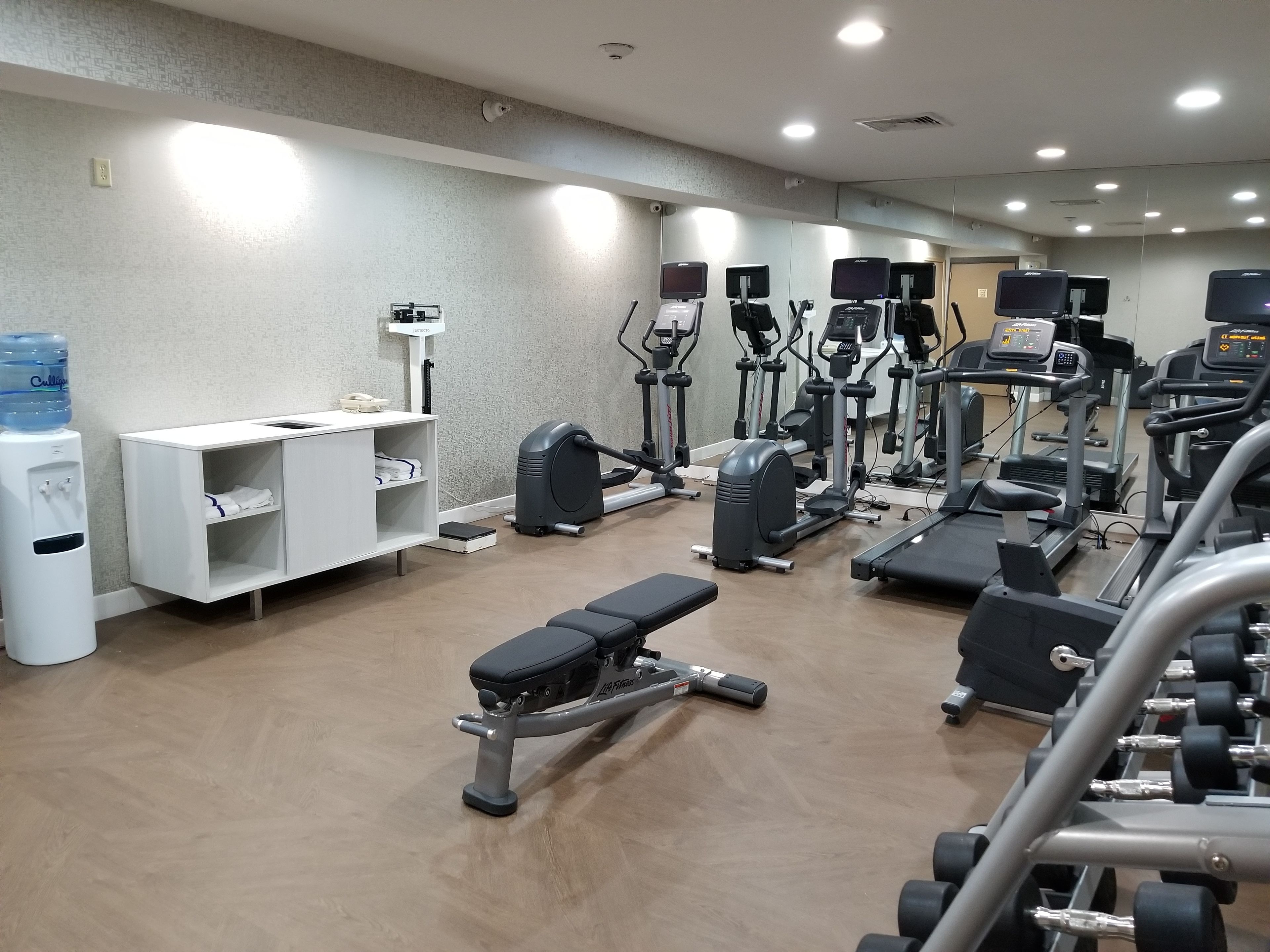Un gimnasio con diferentes equipos de ejercicio, un dispensador de agua y un armario blanco con estantes.