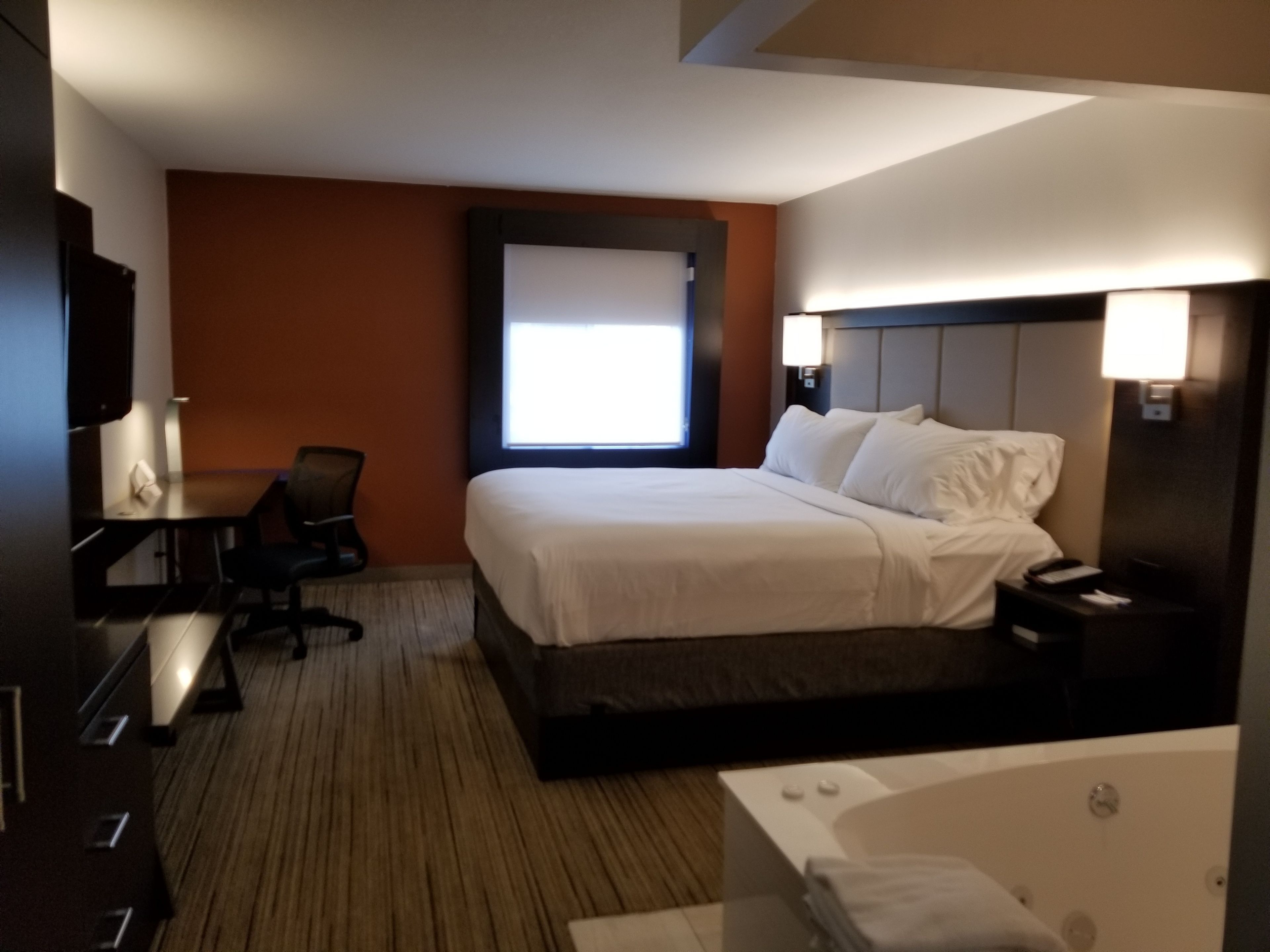 Une chambre d’hôtel avec un grand lit, un bureau, une chaise, une fenêtre, une tête de lit avec des lampes et une baignoire.