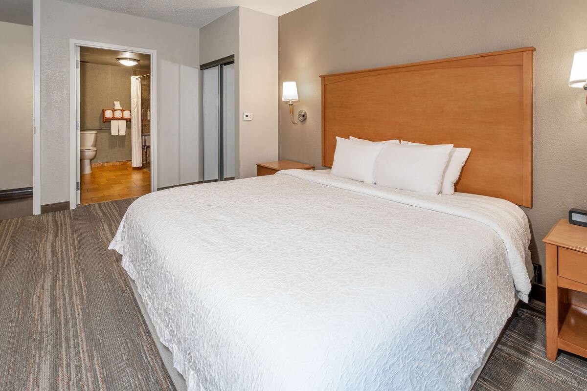 A hotel room with a large bed, a wooden headboard, a nightstand, and a bathroom visible through an open door.
