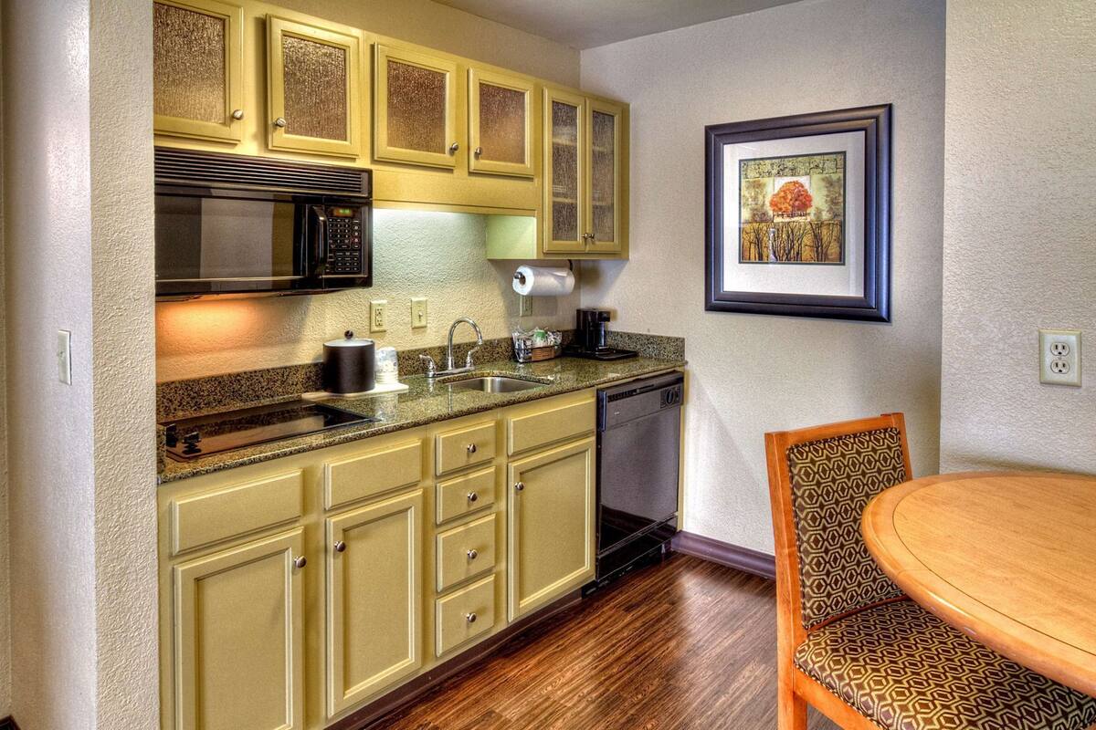 A kitchen with a microwave, sink, and dishwasher, wooden cabinets, and a dining area with a patterned chair.