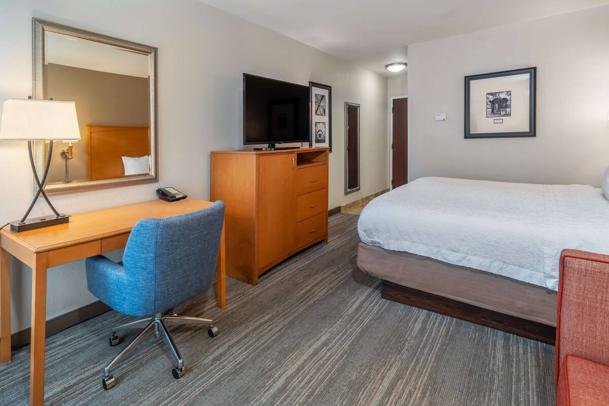 A hotel room with a bed, a desk with a chair, a dresser, a TV, and framed pictures on the wall.