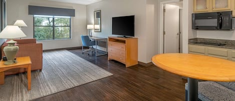 A modern hotel room with a wooden floor, a dining area with a round table, a TV, a desk with a chair, and a kitchenette with a microwave.