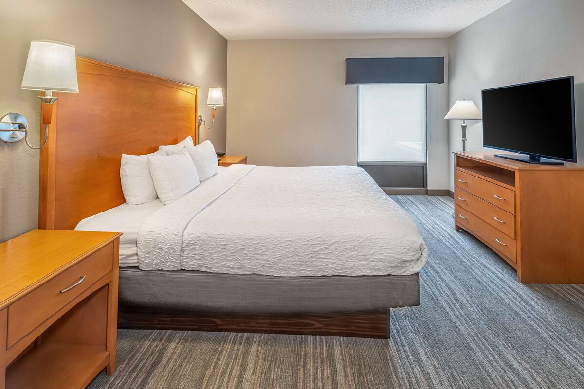 A hotel room with a large bed, wooden headboard, bedside tables, a flat-screen TV, and a window with a view.
