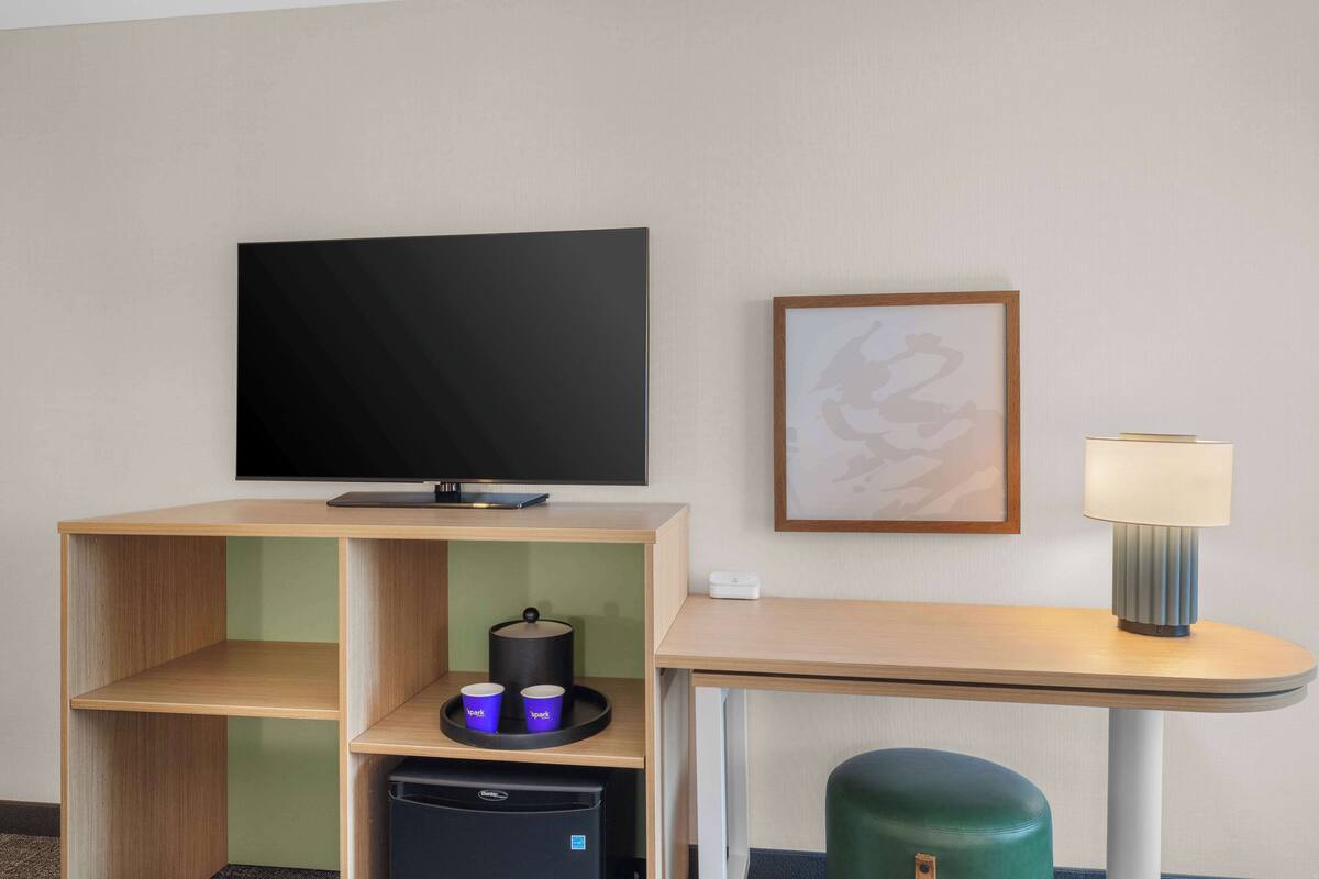 Habitación de hotel con televisor de pantalla plana, una obra de arte enmarco, una lámpara, una mesita, un taburete verde y un minibar.