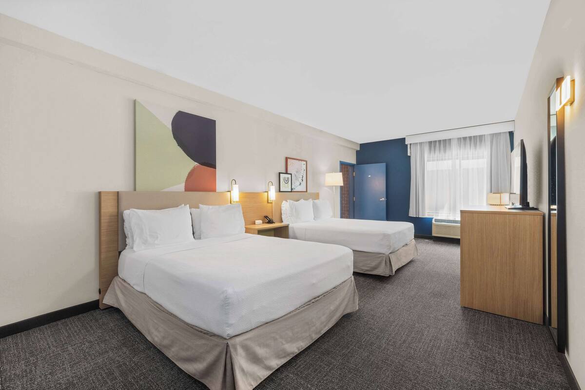 Una habitación de hotel con dos camas, un escritorio, una silla, un televisor y un ventanal con cortinas.