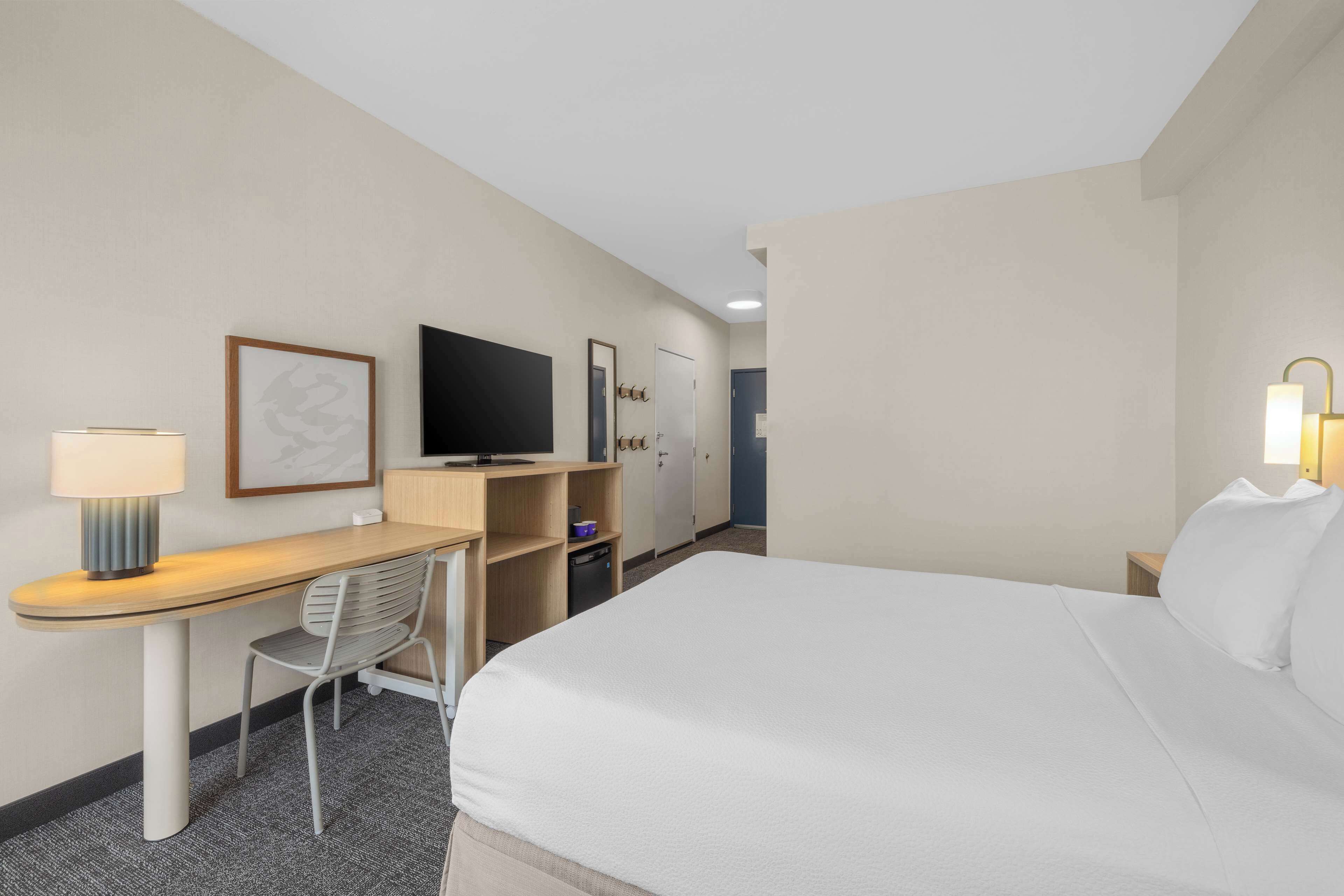 A hotel room with a bed, a desk, a chair, a television, a wardrobe, and a small table with a lamp.
