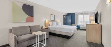A hotel room with a bed, a sofa, a small table, a TV, and a painting on the wall.