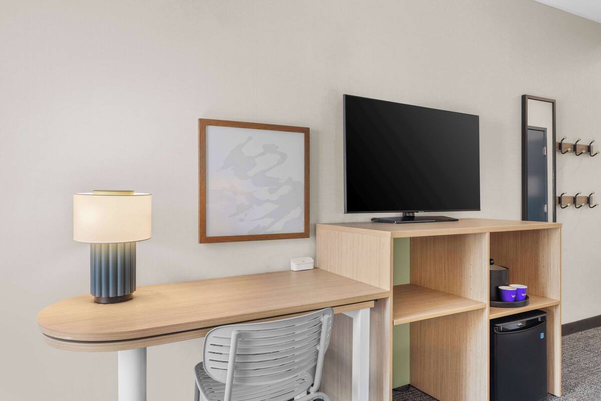 Habitación de hotel con escritorio, silla, televisión, un cuadro enmarcado, espejo y un mueble pequeño.