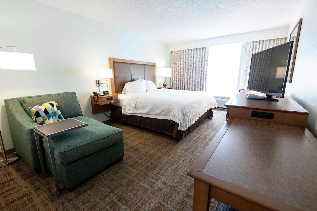 A hotel room with a bed, a green sofa, a small table, a desk, a TV, and a window with curtains.