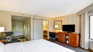 A hotel room with a bed, a desk with a TV, a chair, a desk lamp, and a bathroom visible through an open door.