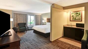 A hotel room with a bed, a desk with a flat-screen TV, a chair, a window with curtains, a painting on the wall, and a kitchenette area.