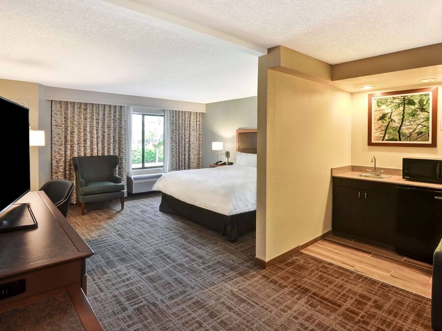 A hotel room with a bed, a desk with a flat-screen TV, a chair, a window with curtains, a painting on the wall, and a kitchenette area.