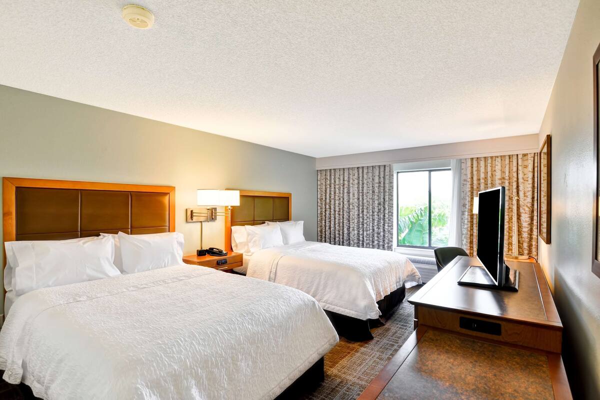 Quarto de hotel com duas camas, uma escrivaninha com televisão, uma lâmpada e uma janela com cortinas.