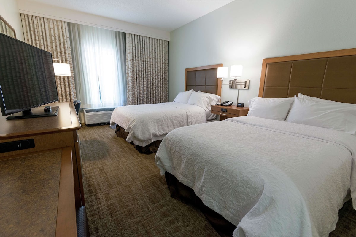 A hotel room with two beds, a TV, a desk, and a window with curtains.