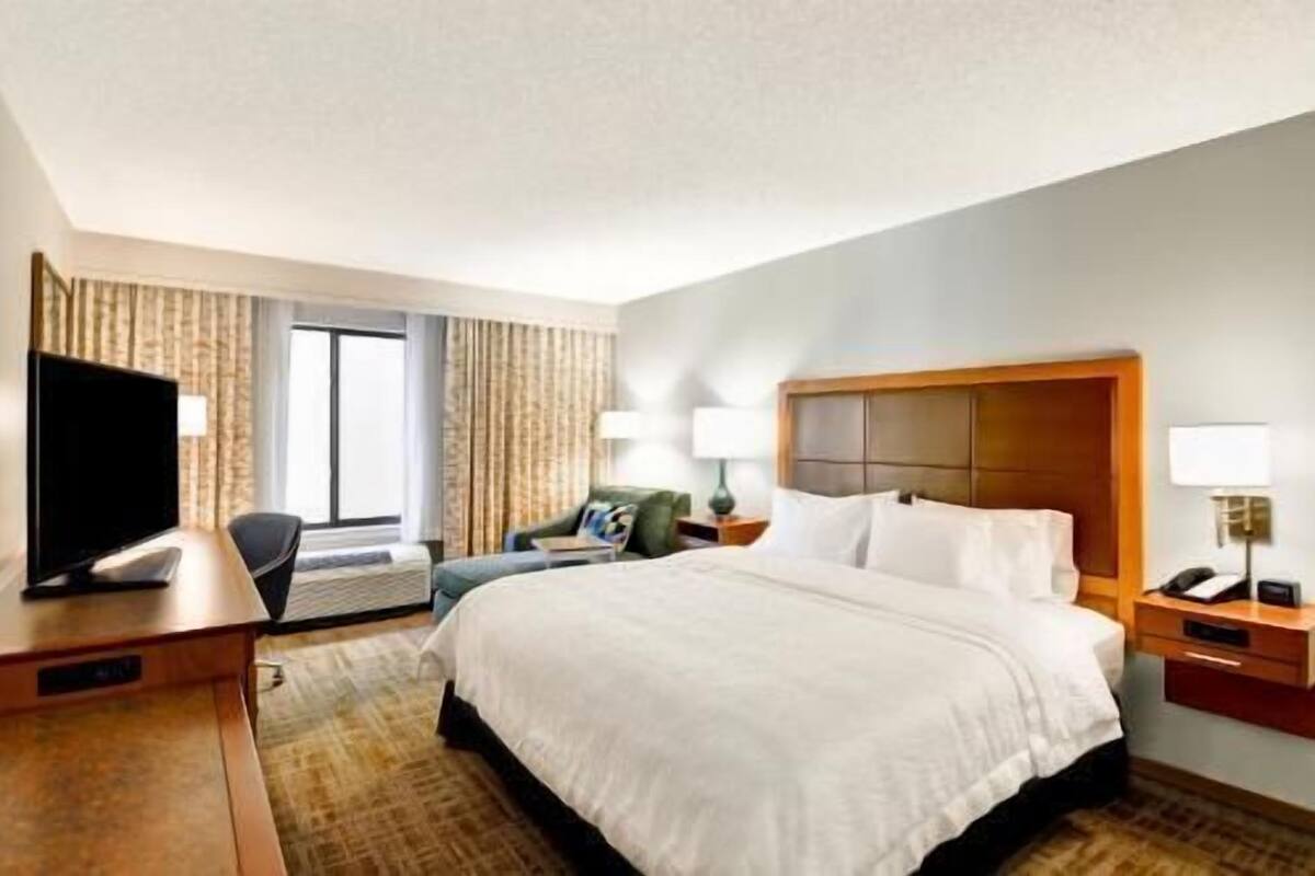 Quarto de hotel com uma cama grande, escrivaninha com televisão de tela plana, uma cadeira e janela com cortinas.
