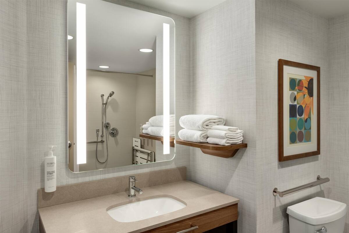 Un baño moderno con un espejo grande, un lavamanos, un inodoro y una ducha. Hay un perchero con toallas dobladas y una obra de arte enmarcada en la pared.