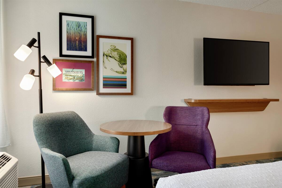 Habitación de hotel con cama, una mesa redonda, dos sillas, televisor y cuadros en la pared.