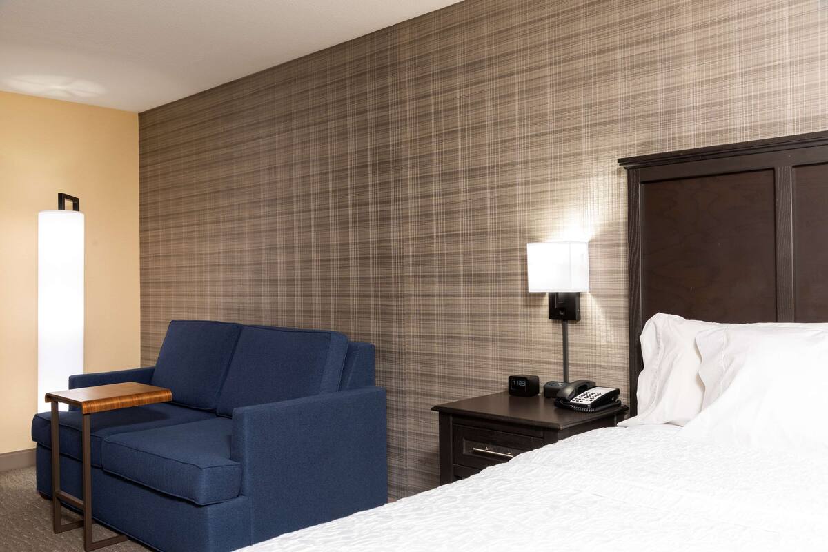 A hotel room with a blue sofa, a wooden side table, a bedside table with a lamp, a phone, and a bed with white bedding.