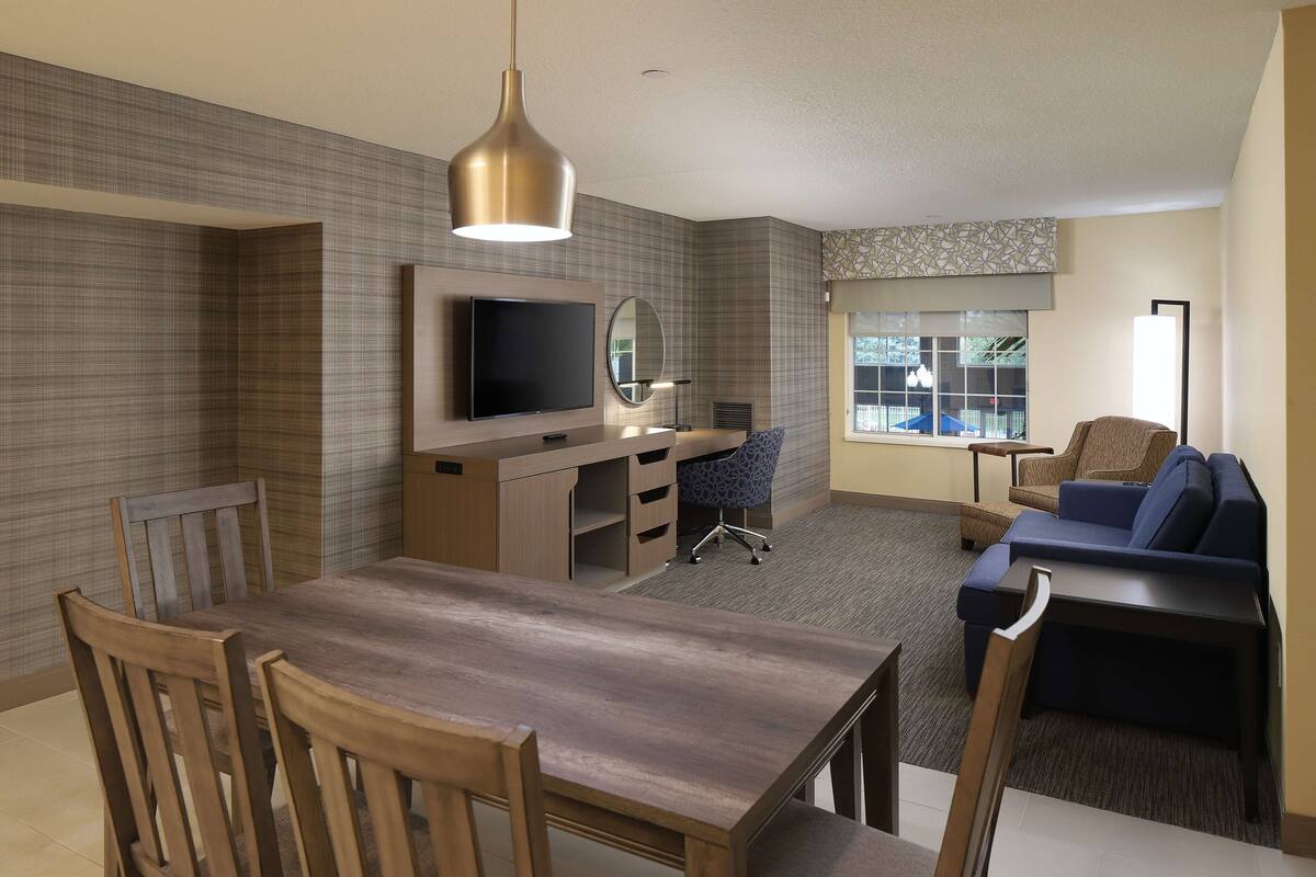 A modern hotel room with a dining area, a sofa, a TV, and a large window.