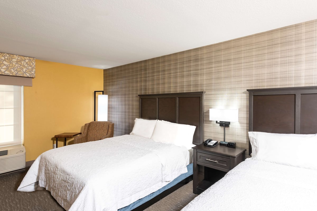 A hotel room with two beds, a nightstand with a lamp, a chair, and a window with blinds.