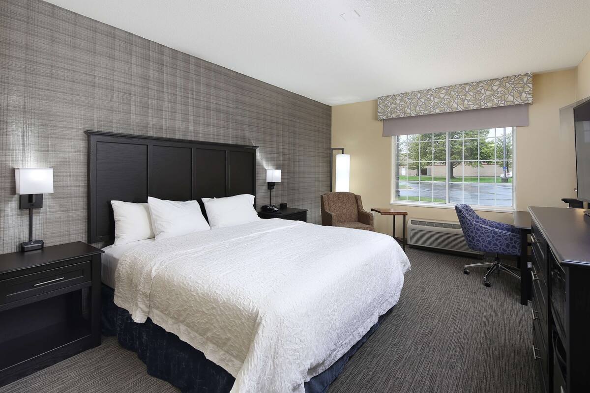 A hotel room with a large bed, a desk, a chair, and a window with a view of the parking lot.