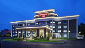 Un hotel Hampton Inn amb un disseny modern, que destaca un llampeu de neó ben visible, diverses plantes i una entrada ben il·luminada.