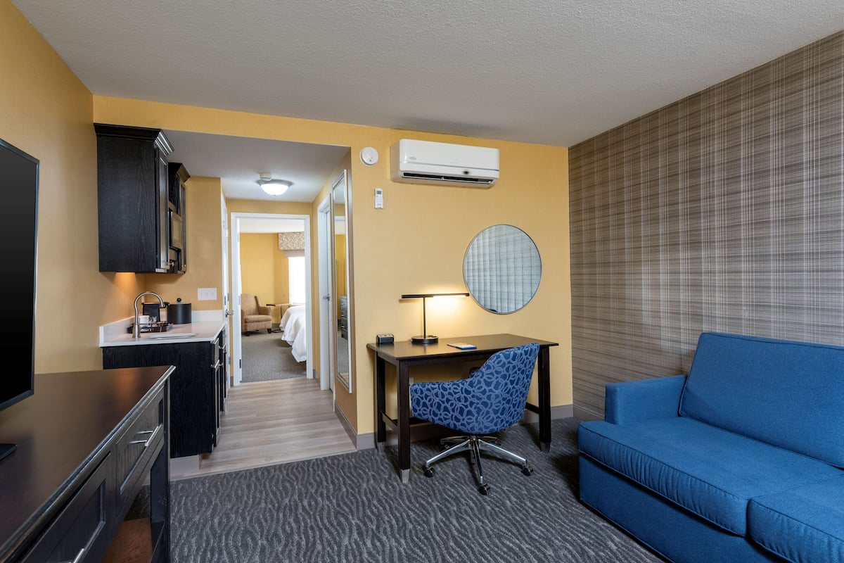 A hotel room with a kitchenette, a desk with a chair, a blue sofa, and a round mirror on the wall.