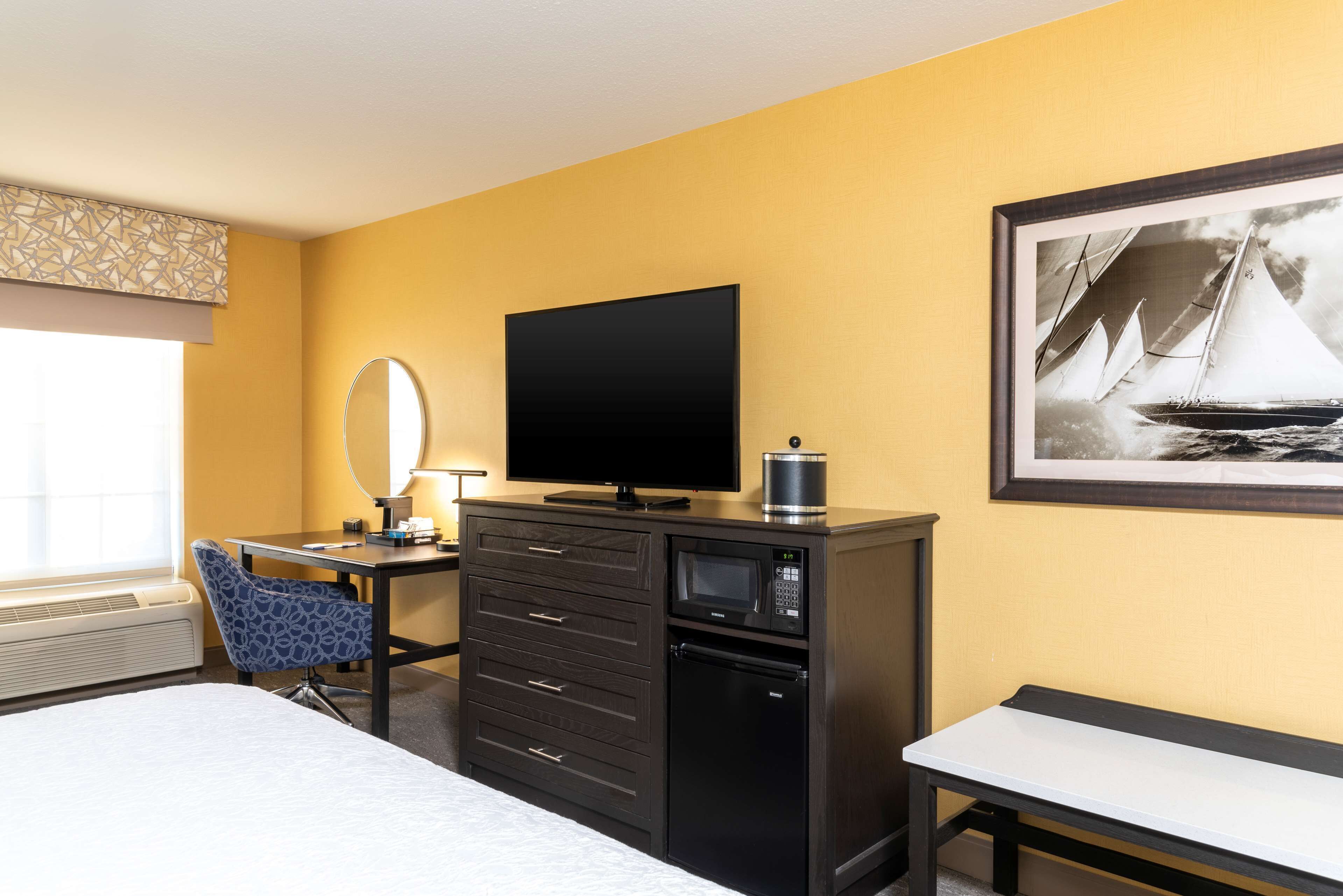 A hotel room with a bed, a desk with a flat-screen TV, a microwave, a refrigerator, a chair, and a framed picture on the wall.