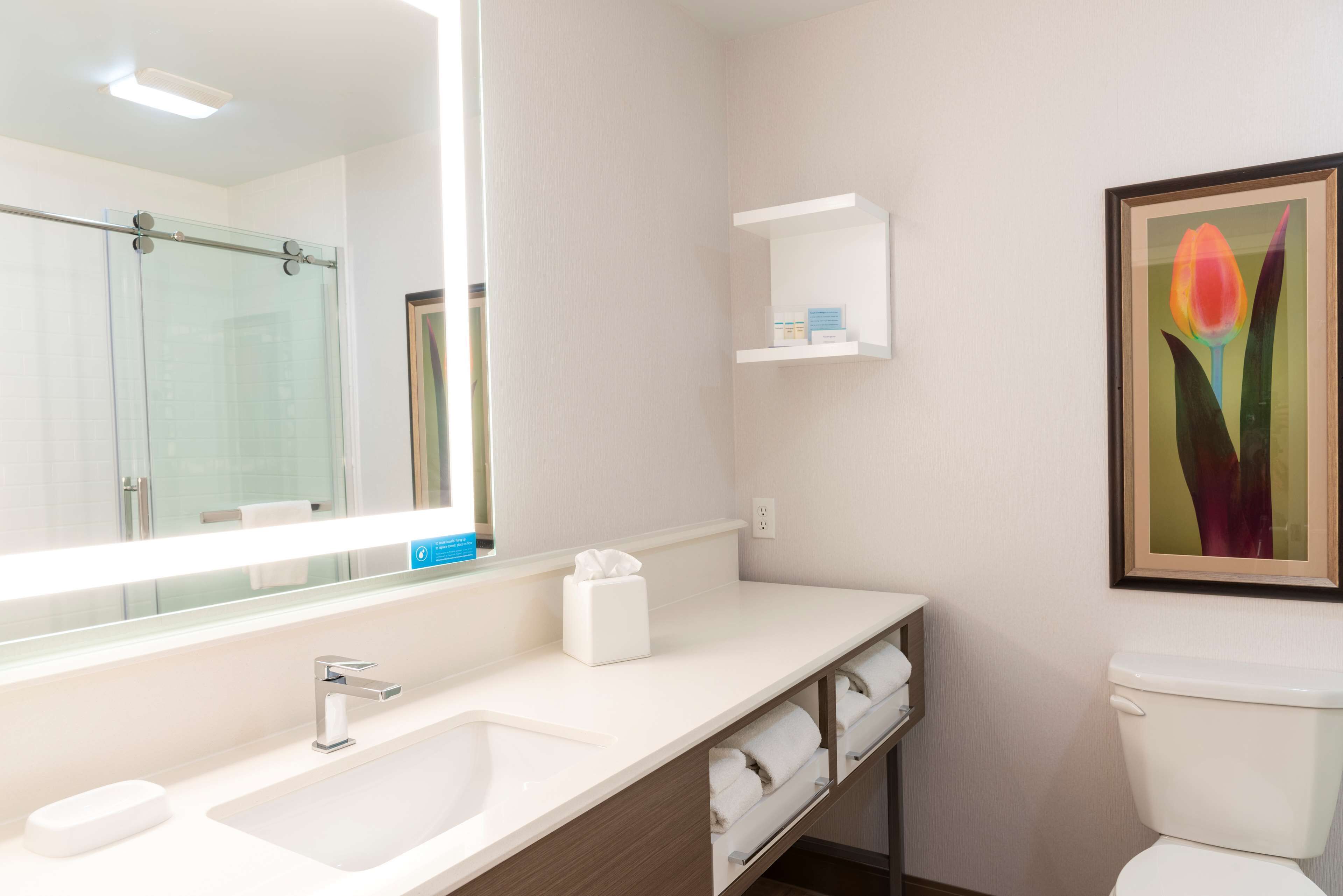 A modern bathroom with a large mirror, a sink, a toilet, and a shelf with toiletries.