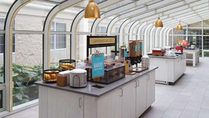 A breakfast bar with a bakery display, a sign indicating breakfast starts here, and a view of the outdoors through a glass ceiling.