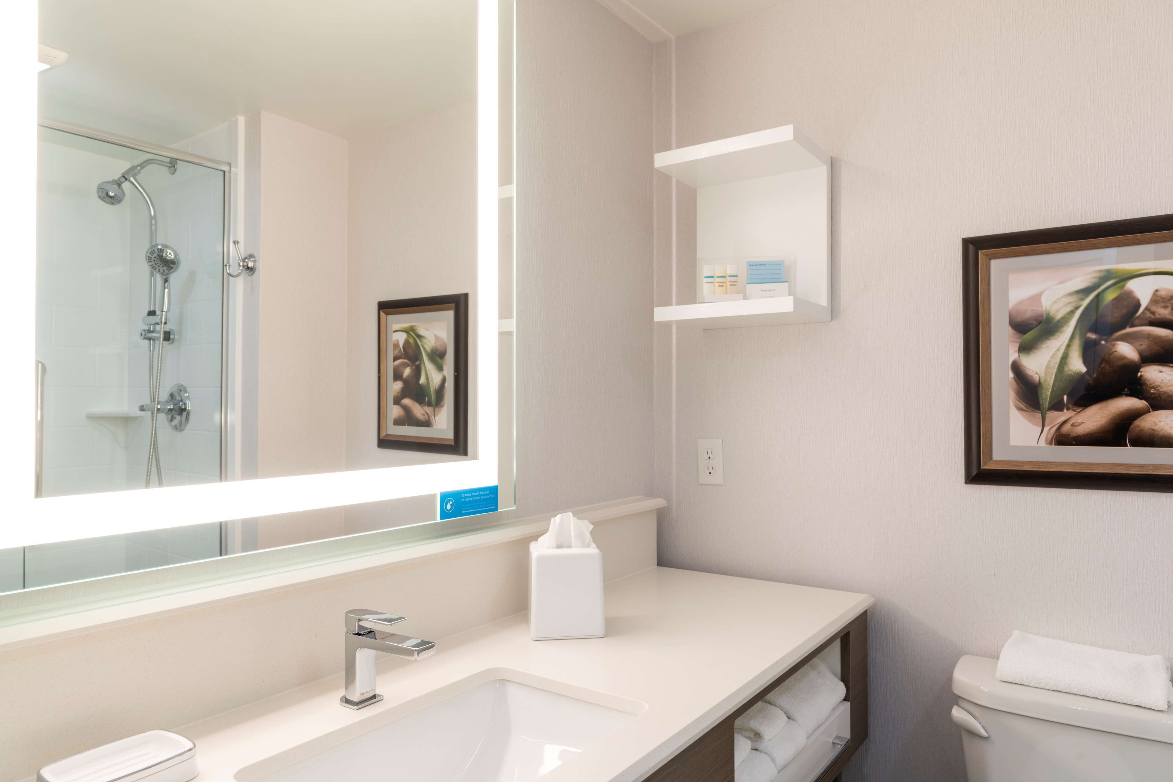 A modern bathroom with a large mirror, a sink, a toilet, and a shower. There are framed pictures on the wall and a small shelf with toiletries.