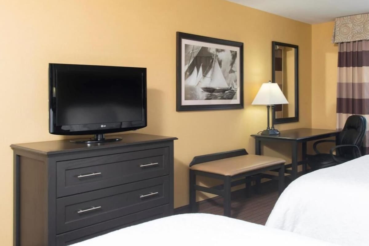 A hotel room with a bed, a TV on a dresser, a desk with a chair, a lamp, and a framed picture on the wall.