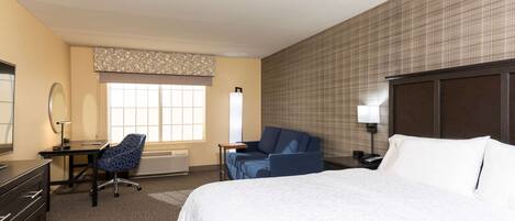 Una habitación de hotel con cama, escritorio y silla, mesita de noche con lámpara, y ventana con persianas.