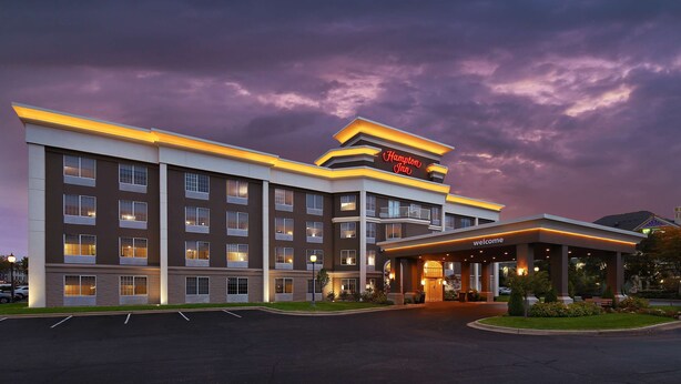 A Hampton Inn hotel with a modern design, featuring multiple floors, a well-lit entrance, and a parking lot.