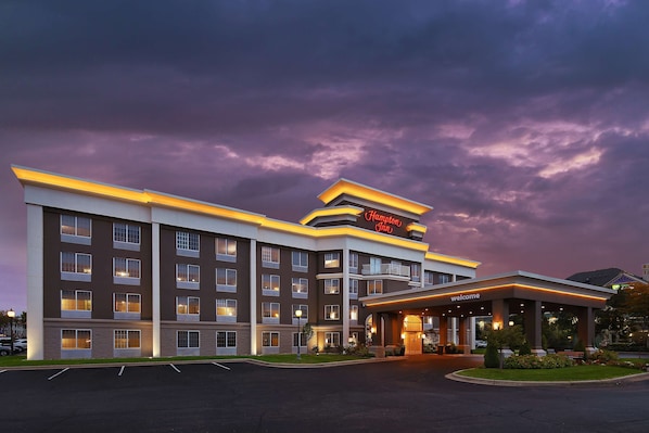 A Hampton Inn hotel with a modern design, featuring multiple floors, a well-lit entrance, and a parking lot.