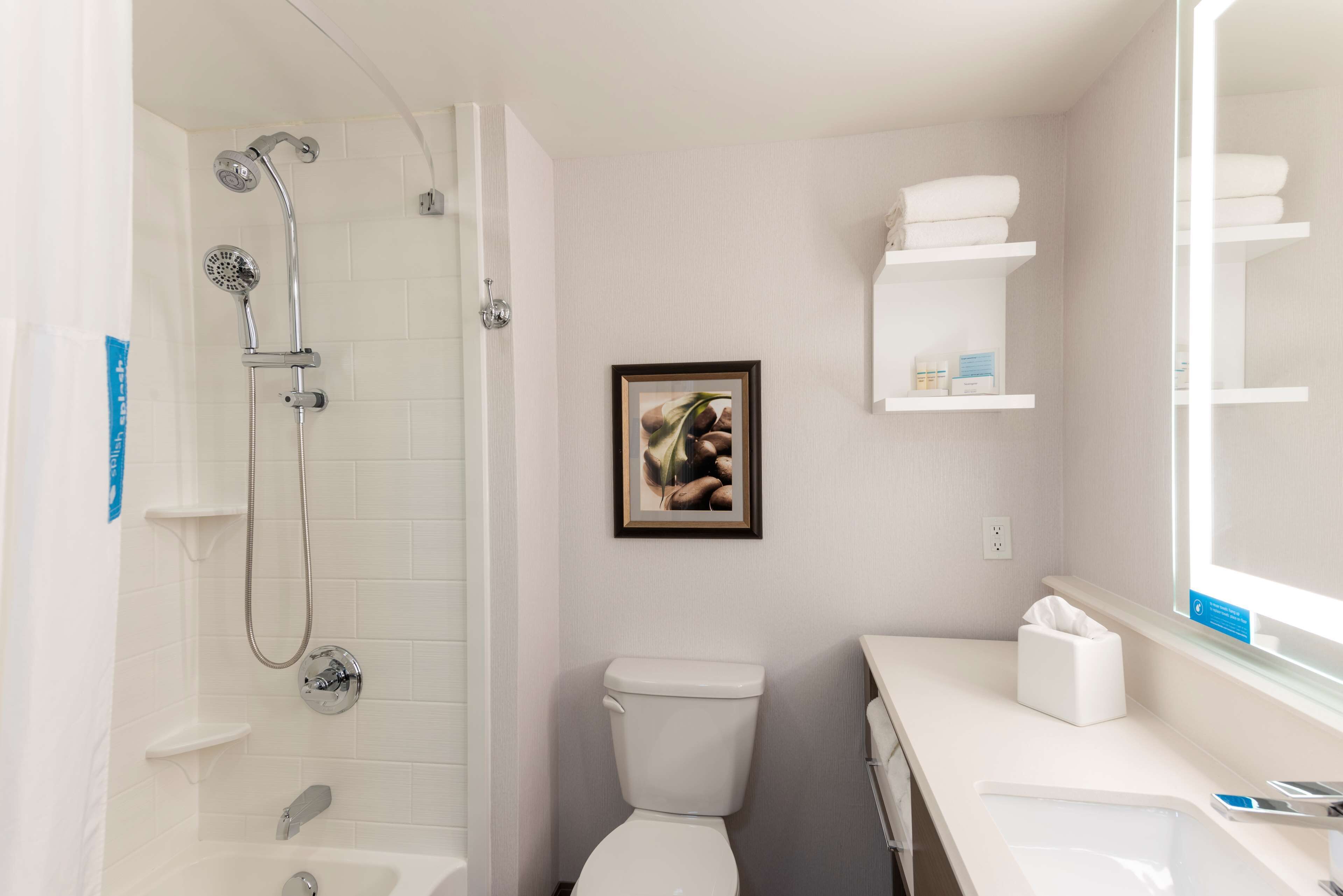 A bathroom with a shower, toilet, and sink. There is a framed picture on the wall and shelves with towels.