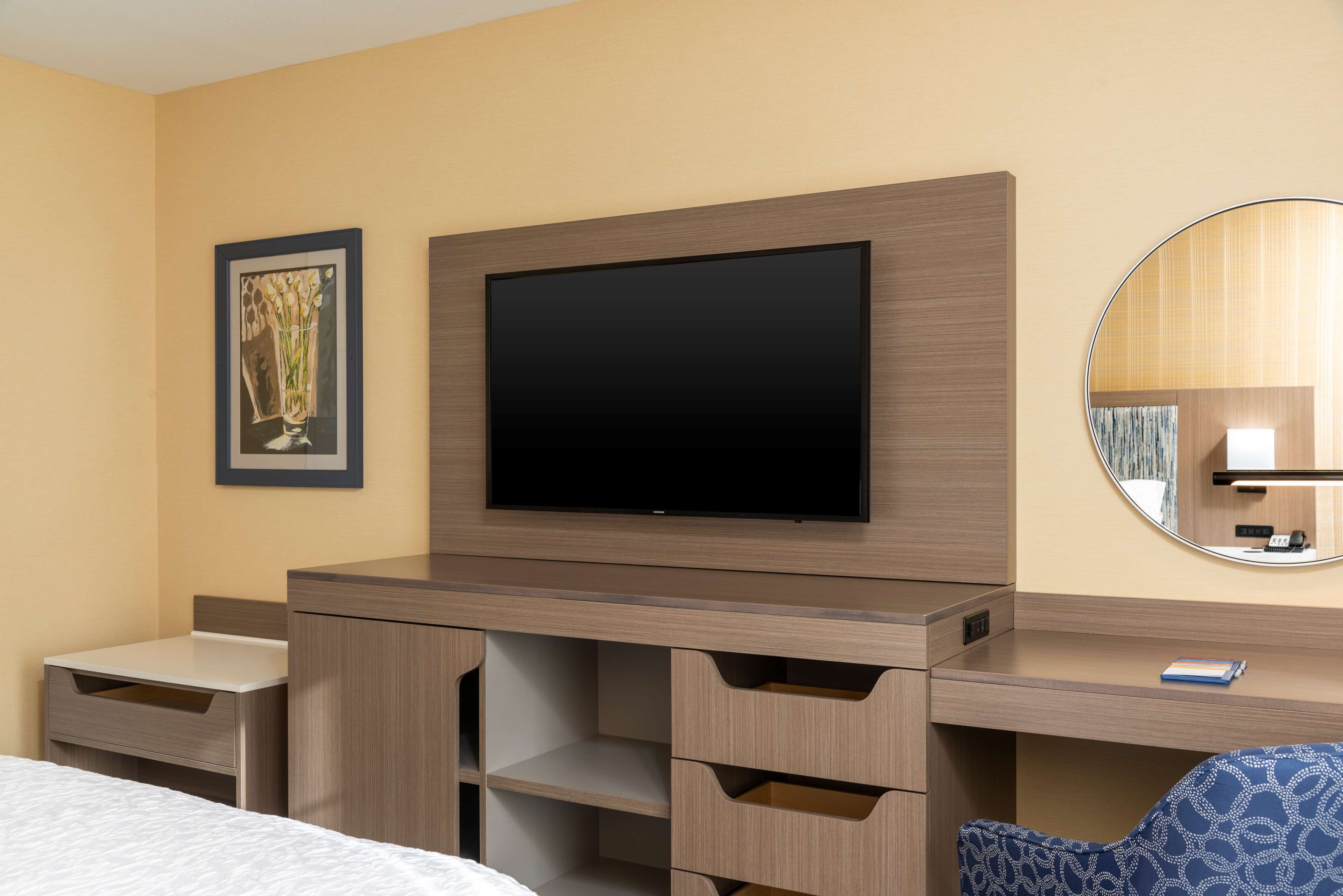 A hotel room with a bed, a desk with drawers, a flat-screen TV, a framed picture, and a round mirror.
