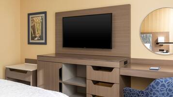 A hotel room with a bed, a desk with drawers, a flat-screen TV, a framed picture, and a round mirror.