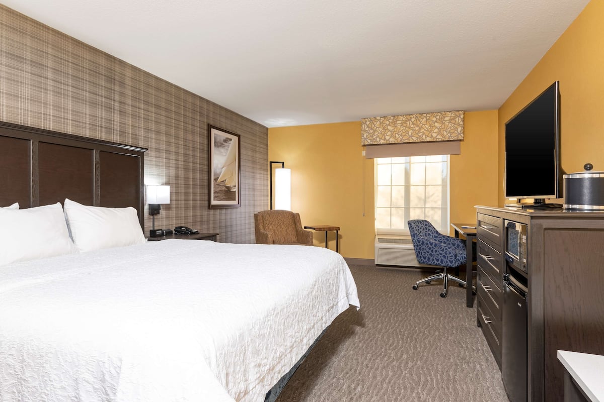 A hotel room with a large bed, a desk with a computer, a chair, a television, and a window with blinds.