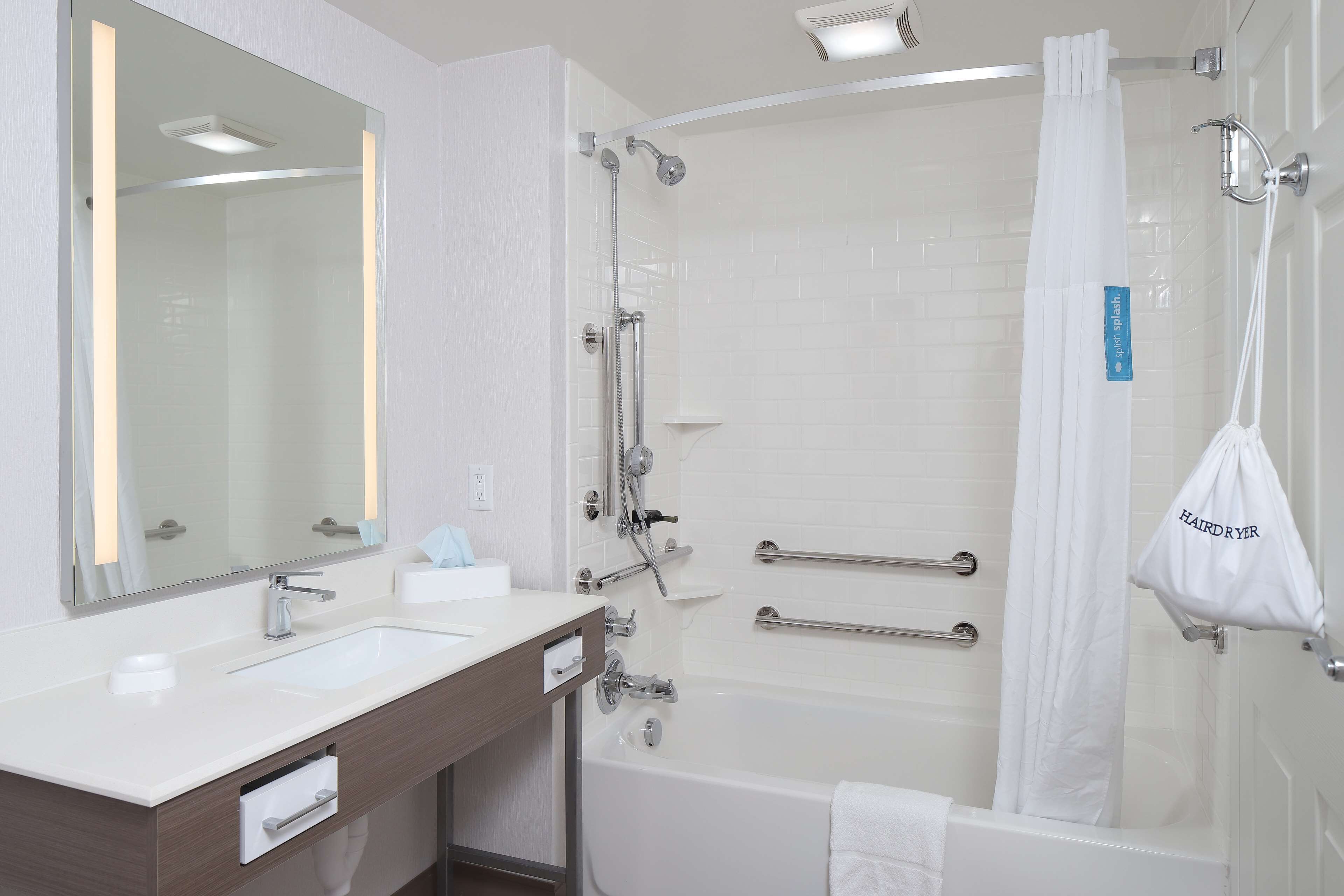 A modern bathroom with a white sink, mirror, and bathtub. There are grab bars in the shower and a white bag labeled "HAIRD RYER" hanging on the wall.