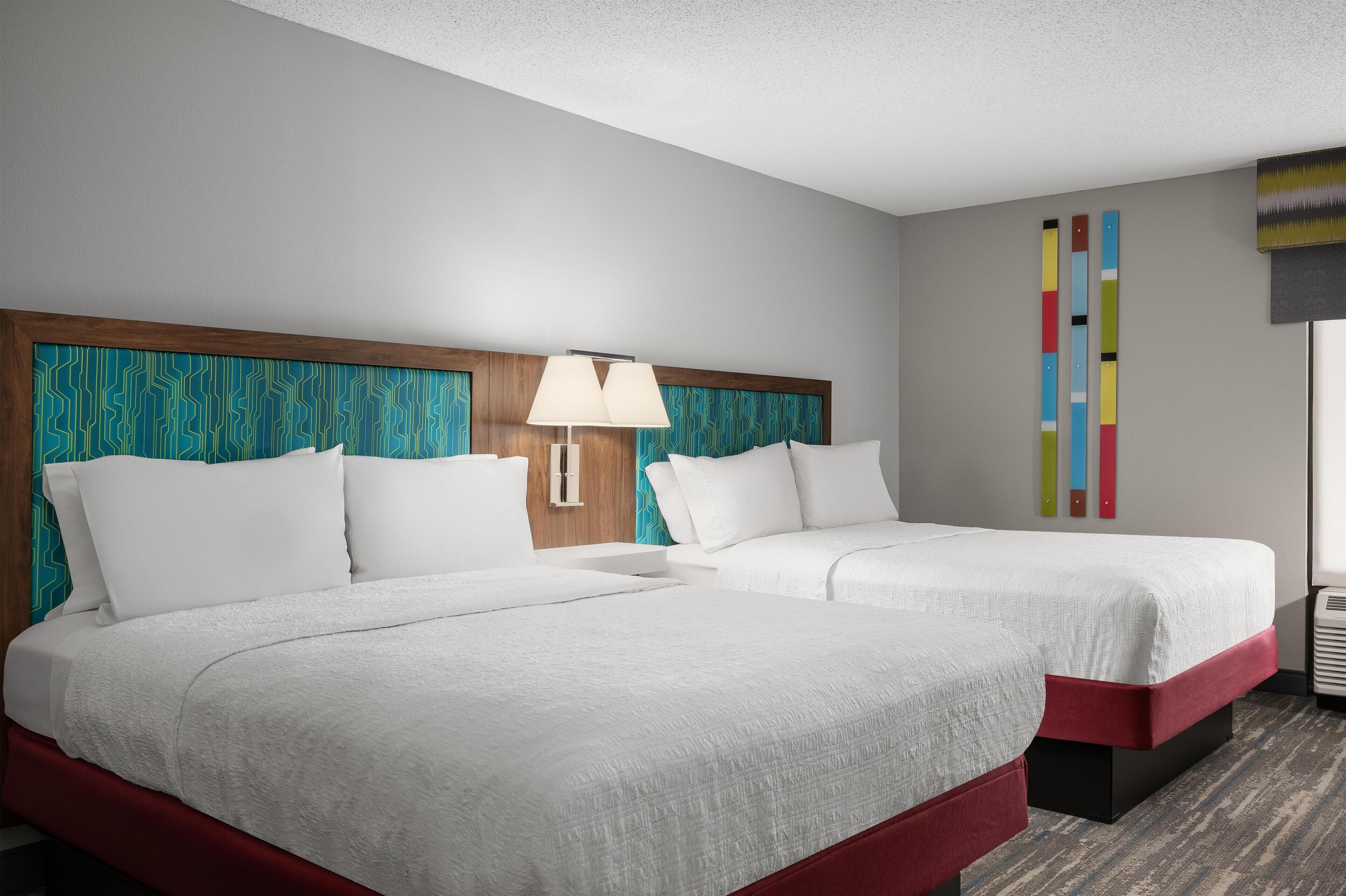 A hotel room with two beds, a wooden headboard, a wall-mounted lamp, and a colorful abstract art piece.