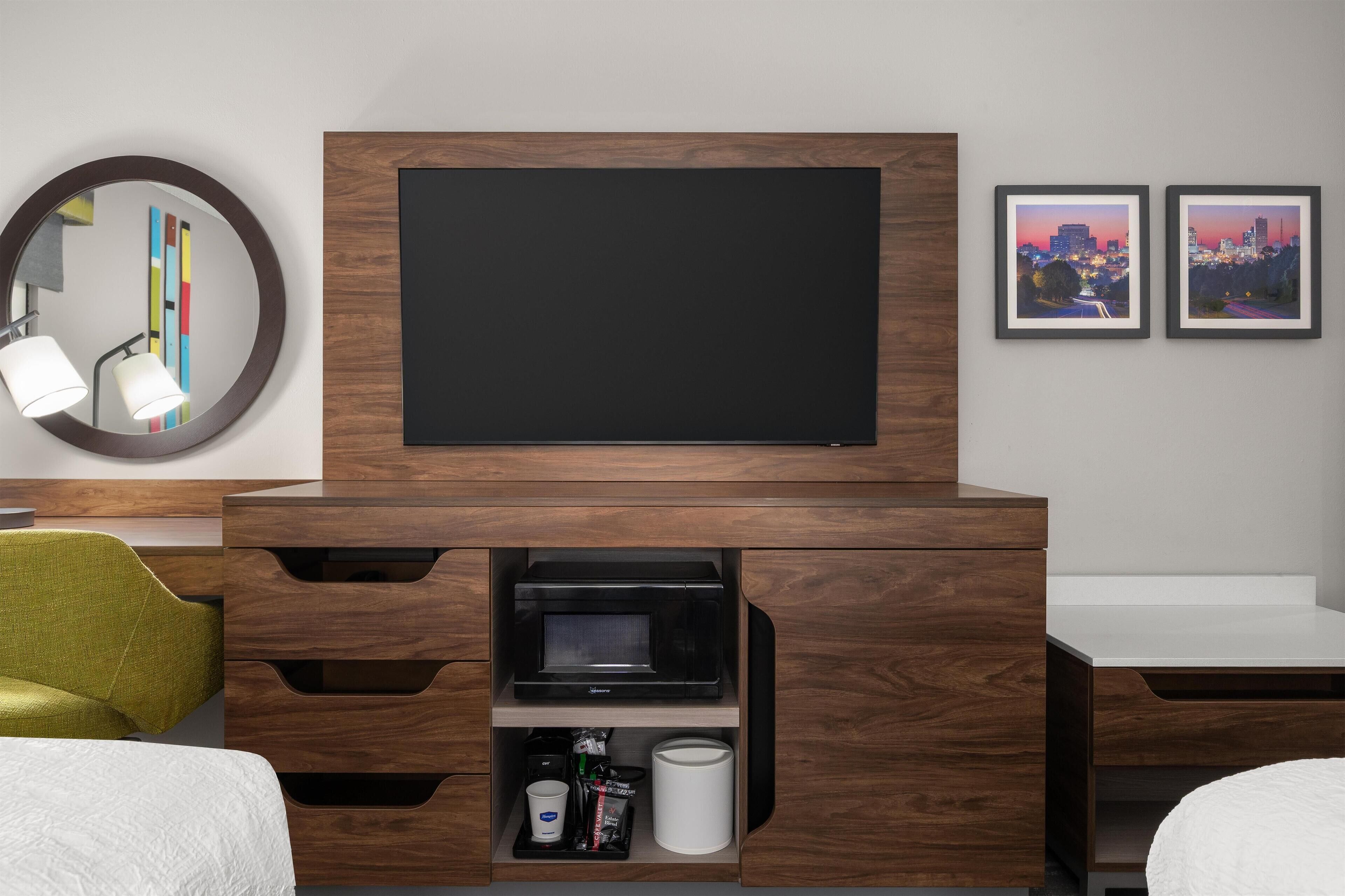 A modern hotel room with a flat-screen TV mounted on a wooden cabinet, a round mirror with a lamp, and two framed cityscape pictures on the wall.