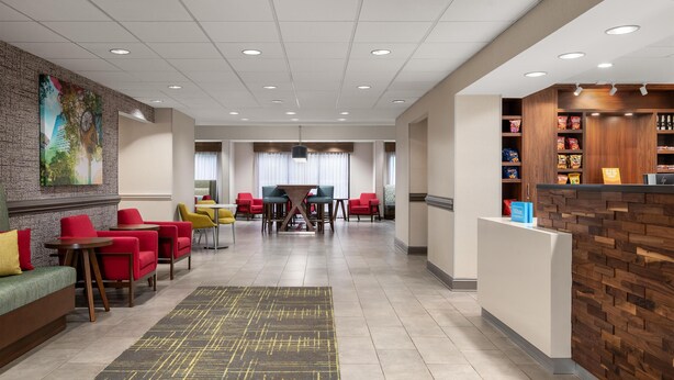 A hotel lobby with a reception desk, seating area with red and yellow chairs, a large painting on the wall, and a wooden reception desk with snacks.