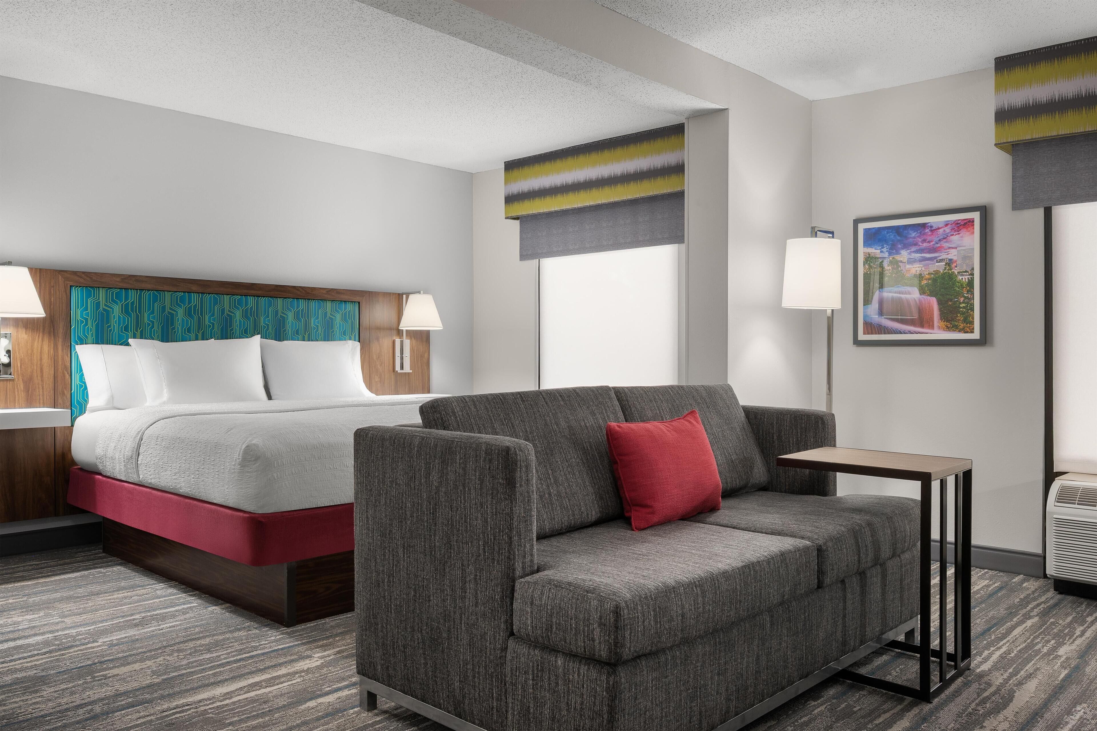 A hotel room with a bed, a sofa, a small table, a lamp, and a picture on the wall.