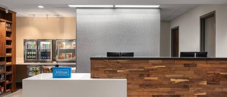 A hotel reception area with a wooden reception desk, a minibar, and a seating area with a view of the lobby.