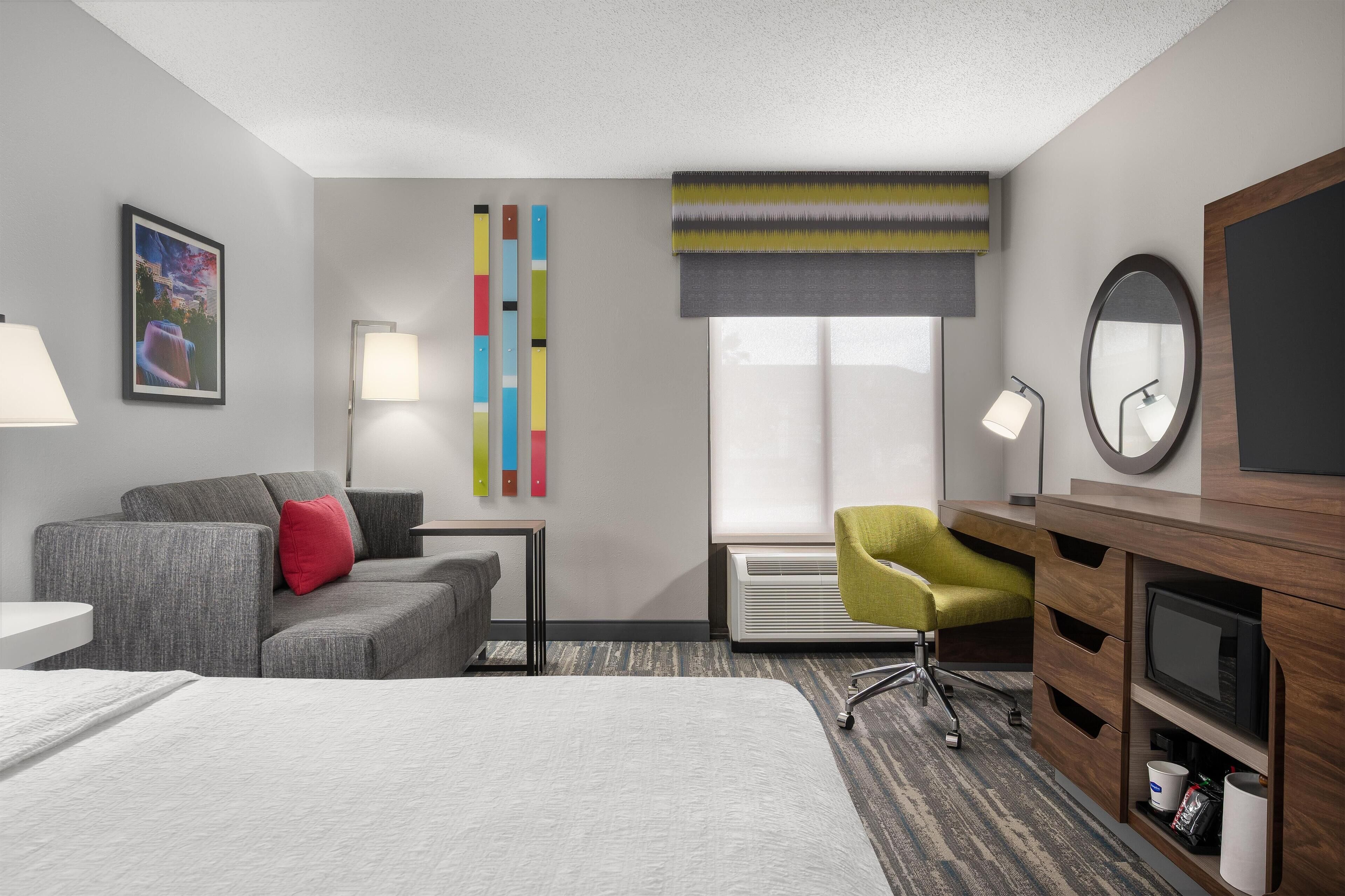 Habitación de hotel con una cama, un sofá gris, un sillón verde, un escritorio con lámpara, un espejo y un televisor.
