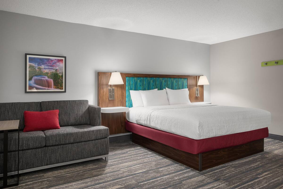 A hotel room with a bed, a sofa, a nightstand, and a wall-mounted lamp.
